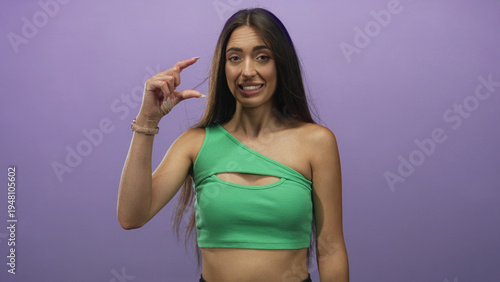 Fotografie Woman in green crop top making a small pinch gesture with thumb and index finger at eye level in studio with purple backdrop; playful