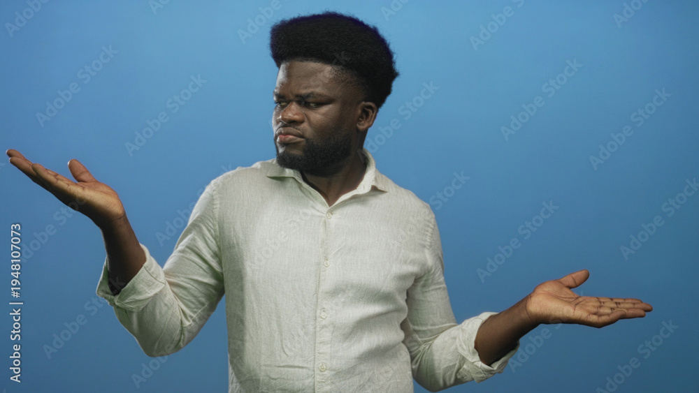 Fototapeta premium Man shrugging with palms up and furrowed brow against a blue studio wall, wearing a light linen shirt with bare hands visible as he gestures unsurely; uncertainty.