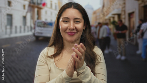 Fotografija Young hispanic woman clapping hands on busy city street during daytime with blurred pedestrians; celebration joy