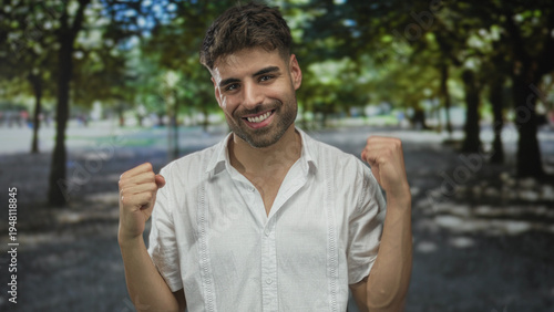 Smiling young hispanic man points finger to camera in sunlit green park clearing amid leafy canopy; confidence.