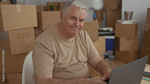 Принти на полотні Man senior typing on laptop at a wooden table surrounded by stacked cardboard moving boxes and a drill in a building; planning determination