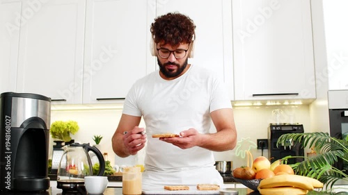 Arabian man dances with headphones in kitchen spreading peanut butter on toast for breakfast. Young guy feels happy preparing quick morning meal with coffee aroma and a tasty healthy routine at home.