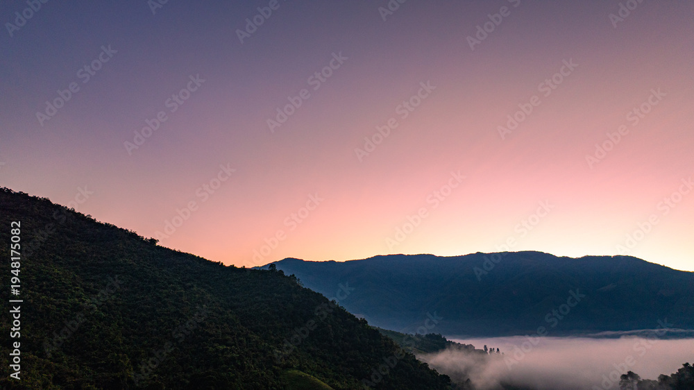 Fototapeta premium Scenic drone view of a misty mountain valley at sunrise in Sapan Village, Nan Thailand. A small village glows beneath drifting fog and soft golden light, showcasing a peaceful countryside travel