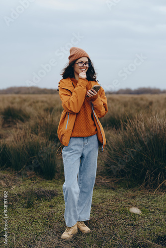 A smiling woman lifestyle enjoys nature outdoors wearing orange jacket and beanie, holding smartphone in hand. Casual autumn outfit with jeans and glasses in a natural grassy field setting.