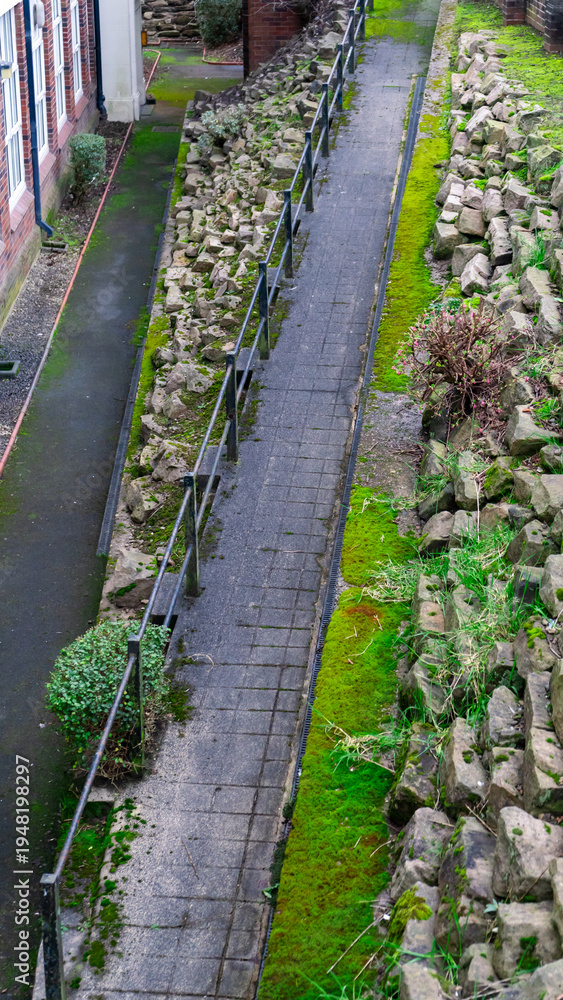 Obraz premium A ginnel a narrow pedestrian passage or alleyway between buildings. The path, paved with cobblestones or slabs, is flanked by stone boulders. View from above