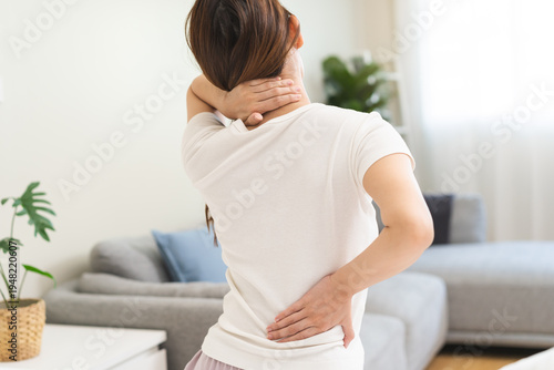 Fotografie Pain body muscles stiff problem, asian young woman painful back, neck ache from work hand holding massaging rubbing shoulder hurt, sore sitting on bed while wake up at home