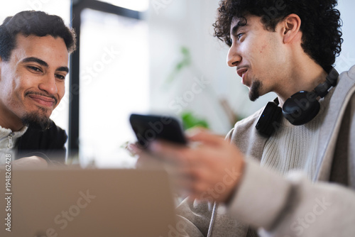 Two young men are interacting with a mobile device and a laptop, sharing information and collaborating on a project.
