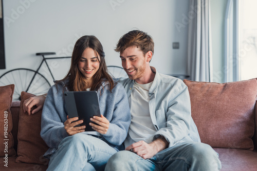 Happy young couple relaxing on cozy couch, using computer tablet together, smiling beautiful woman and man hugging, looking at device screen, watching movie or video, shopping online, browsing apps
