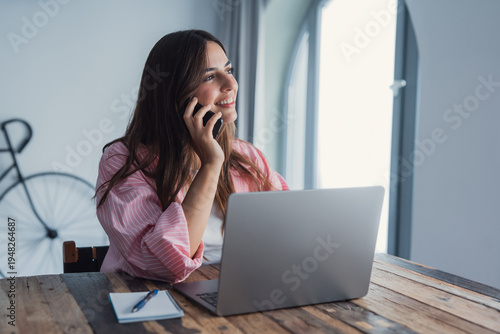 Smiling young lady working at home office desk by laptop engaged in phone conversation with client give remote consultation. Caucasian woman employee make business call from workplace distracted