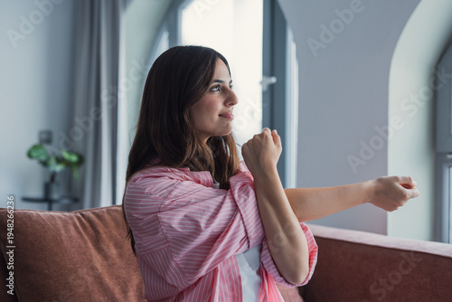 Photography Young woman sitting on a sofa in her living room performing a gentle arm stretch