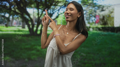 Wallpaper Mural Thai woman points index fingers upward while smiling, wearing a white sundress among trees and grass in a forest; playful confidence. Torontodigital.ca