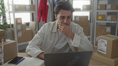 Photos Man at laptop with hand on chin, surrounded by shipping boxes on desk in building workspace; concern small business ecommerce