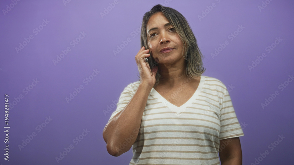 Fototapeta premium Woman holding smartphone to ear while checking messages and then smiling in studio purple backdrop; casual conversation content.