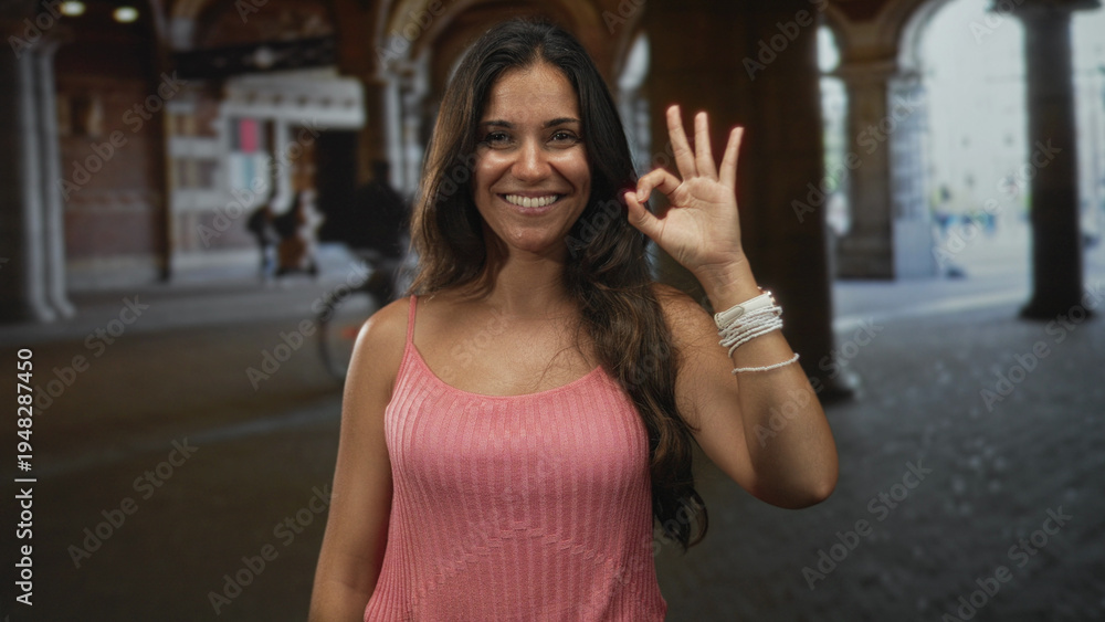 Fototapeta premium Woman showing ok sign with right hand in building archway wearing pink top and bracelet; joy approval.