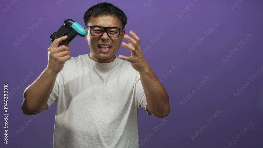 Fototapeta premium Man wearing glasses and white t shirt grimacing holds blue and black game controller and raises hand against purple studio wall; frustration.