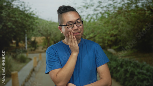 Man with hand on cheek smiling in park pathway surrounded by green trees and shrubs; calm reflection.