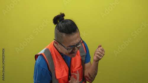 Cuadro en lienzo Man wearing an orange reflective vest clenches fist in a studio against a yellow wall with closed eyes and glasses; triumph