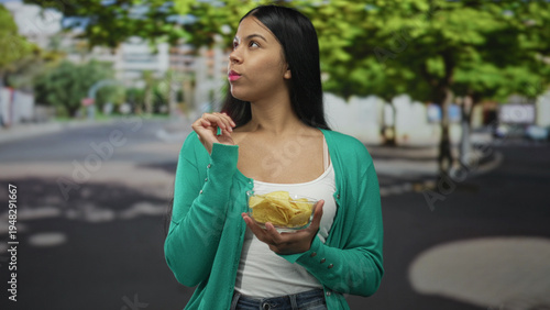 Canvas Print Latin woman holds chip to lips with a bowl of potato chips in hand on a city street; craving satisfaction