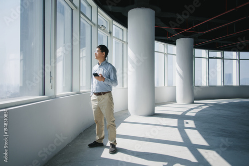 Photos Businessman in office standing by window holding smartphone while gazing into th