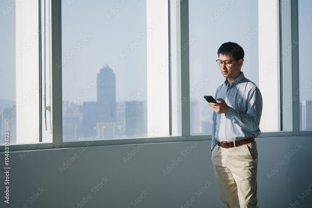 Fototapeta premium Man in office standing by window holding smartphone while looking at screen