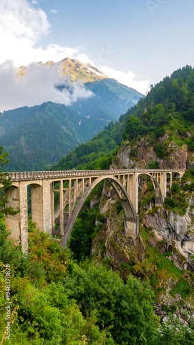 Arched bridge spans a valley of lush green foliage and mountainous terrain under a partly cloudy sky
