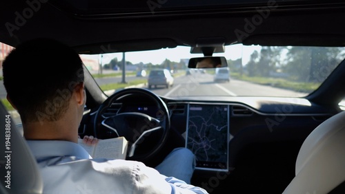 Male businessperson reading book during riding on electrical vehicle with autopilot at urban road. Successful businessman improving his knowledge while riding an autonomous self driving electric car