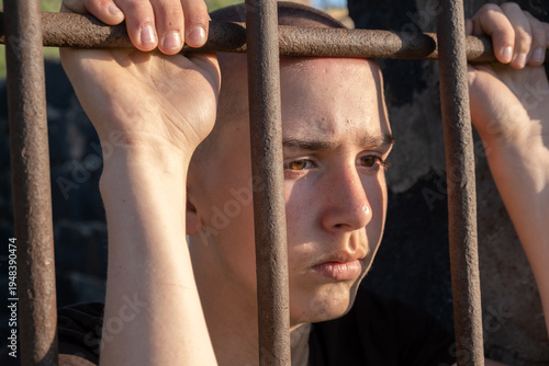 Fotografie Teenage boy with shaved head stands behind prison bars holding the metal grid of a jail window and looking down with sad thoughtful expression
