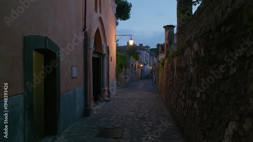 Ravello, Italy. Pan shot of Via San Giovanni del Toro in Ravello at dusk, with warm glowing street lanterns, cobblestone path, terracotta facades and ancient stone walls receding into the distance.