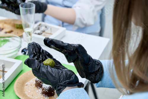 Hands in black nitrile gloves making green matcha truffle ball during culinary master class with chocolate desserts and coconut flakes on table in professional pastry workshop or home kitchen.