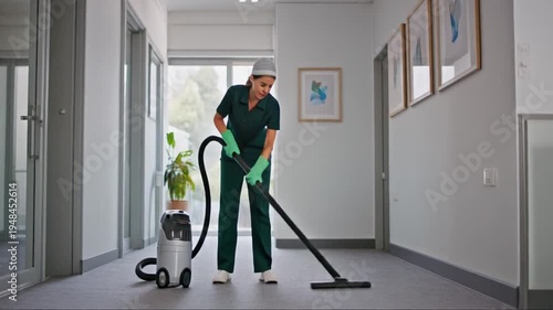 A man in a green uniform vacuuming a modern office hallway with glass doors and framed pictures on the wall