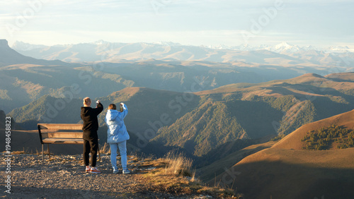 Fotografie Two women take photos of the stunning mountain landscape on their phones early in the morning