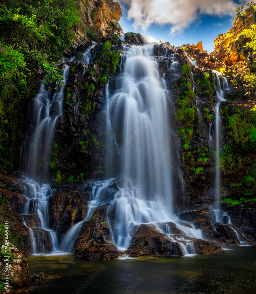 Obraz premium Large multi-tier tropical waterfall cascading over rocky cliffs into a clear pool surrounded by lush green vegetation.