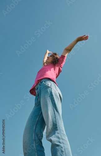 Cuadro en lienzo Beautiful carefree young woman with curly hair in pink t-shirt raising her hands up on blue sky background low angle shot