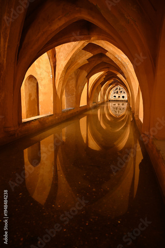 Historic underground corridor with Gothic and Moorish arches reflected in still water inside the Royal Alcazar of Seville, Spain. Atmospheric architectural perspective with symmetry, warm light 