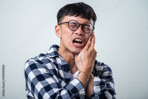 Young Asian man wearing round glasses, plaid shirt, and white tank top holds his cheek and jaw in pain with mouth open against a light gray background