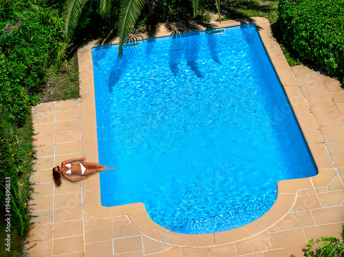 Φωτογραφία Une femme en bikini blanc se détend au bord d’une piscine bleue entourée de verd