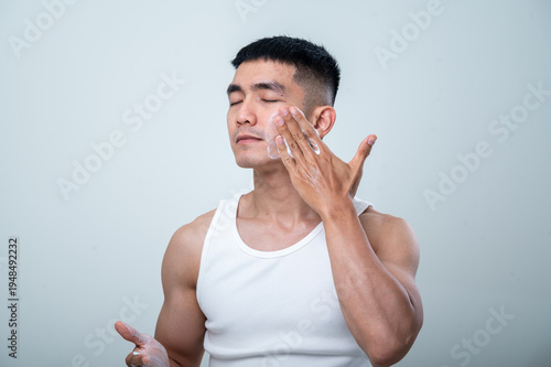 A young Asian man in a white tank top applies facial cleanser to his cheek with his hand, showing a focused expression while performing his skincare routine against a neutral background