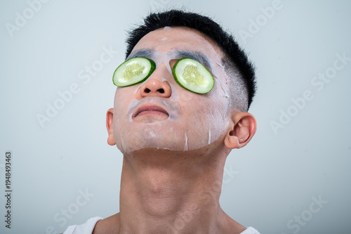 A young Asian man in a white tank top relaxes with cucumber slices over his eyes and a facial sheet mask on his face. He tilts his head back, enjoying a skincare routine