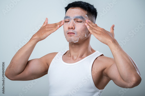 A young Asian man in a white tank top adjusts a facial sheet mask on his face, gently pressing it with his hands. He has his eyes closed, enjoying the skincare routine against a neutral background