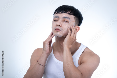 Young Asian man in a white tank top applies a facial sheet mask with both hands against a light gray studio background