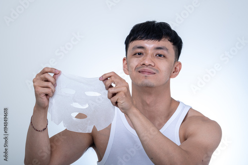 Young Asian man in a white tank top holds a facial sheet mask in both hands while posing against a light gray studio background