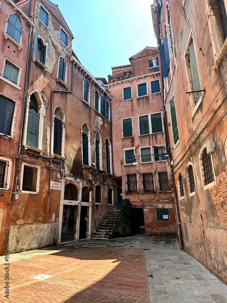 Obraz premium Historic buildings in Venice with faded pink walls and green shutters. Narrow street with pavement and historical architecture create the atmosphere of an old European city.