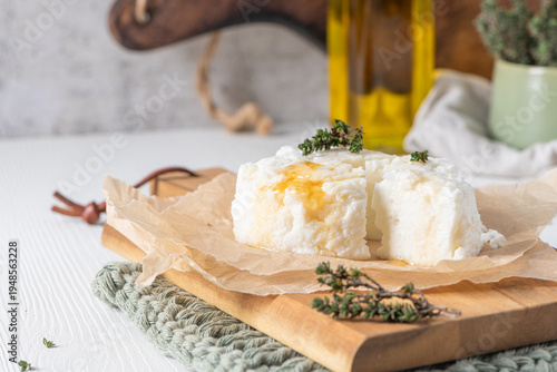 Soft white cheese drizzled with golden honey and topped with fresh thyme on a wooden board with parchment paper in a bright rustic kitchen setting featuring olive oil and greenery in the background
