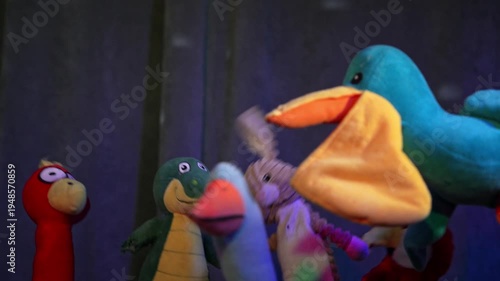 Three colorful bird puppets or toy figurines dance under blue stage lighting next to a glowing yellow lampshade, creating a whimsical theatrical scene.