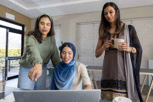 Obraz na płótnie Diverse women in hijab and patterned dress leaning, pointing at laptop in meetin