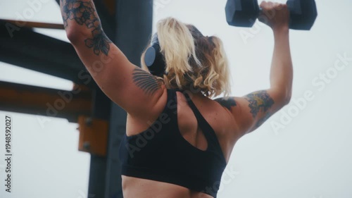 Tattooed Athletic Woman Doing Dumbbell Lateral Raises from Behind Under Graffiti Bridge at Urban Waterfront