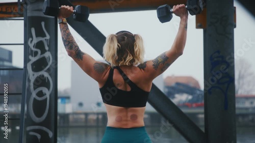 Tattooed Athletic Woman Doing Dumbbell Lateral Raises from Behind Under Graffiti Bridge at Urban Waterfront