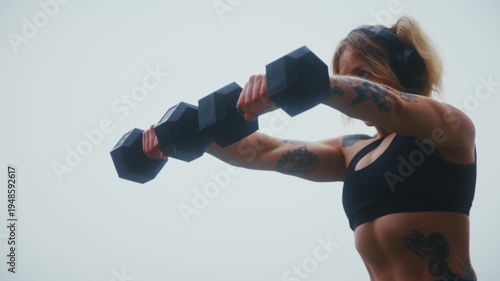 Tattooed Athletic Woman Doing Dumbbell Front Raise with Headphones at Industrial Harbor Waterfront