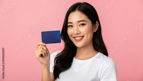 Credit Card in Hand: An Asian woman with radiant smile, her gaze direct, proudly displays a credit card against a backdrop of soft blush.