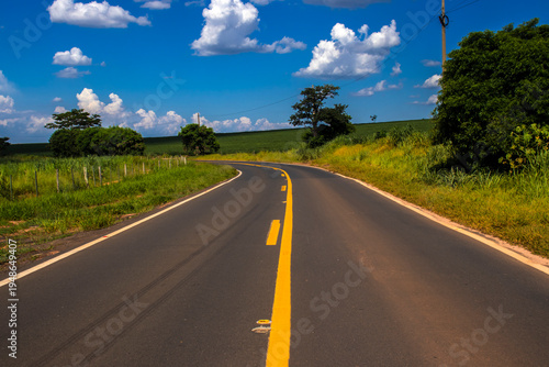 Asphalt road with yellow lines curves through the vibrant green landscape of rural Brazil under a blue sky. A concept of journey, travel, and perspective in South America's countryside.